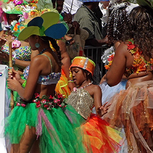 Women with colorful costumes dance in the street, a young girl with a metalic silver dress with orange, red, and yellow skirt and hat dances in the center. 