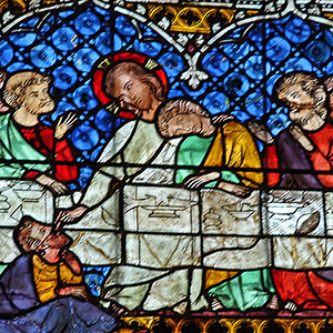 Last Supper in Stained Glass Jesus sits in the center of the table with his left arm resting on the shoulder of one disciple, while, with his right arm, he feeds another disciple who is crouched in front of the table.