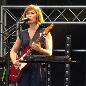 Mina Tindle up on stage singing "Pas les saisons", she is squinting into the lights to see the crowd, holding her guitar, with her mouth near the standing microphone.