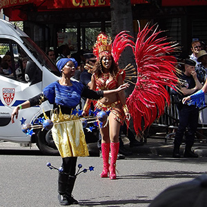 A woman with arms outstretched, wearing a blue shirt and yellow skirt, dances in front of a woman with a giant red feather costume.