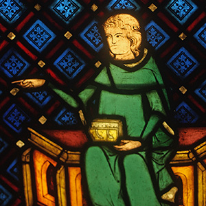 Young cleric in green A young man in a green robe sits with a box in his lap. He is pointing to the left. Behind him is a pattern of blue diamond-shaped squares with red borders.