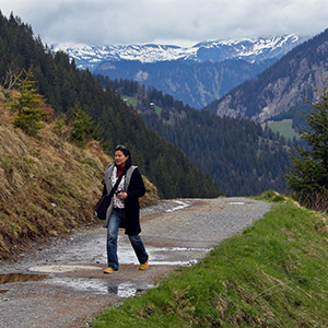 Jeri is walking on a trail with mountains and forests behind her.