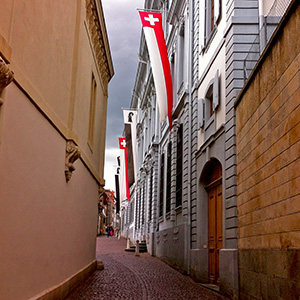 A narrow alley leads toward a blue-grey building with streaming banners of Basel and Switzerland