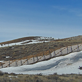 Drifts of snow lie along the snow fence on a sagebrush-covered slope
