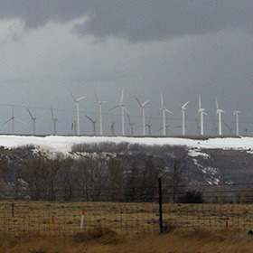 A long line of wind turbines loom above the white snow, below the dark and threatening clouds.