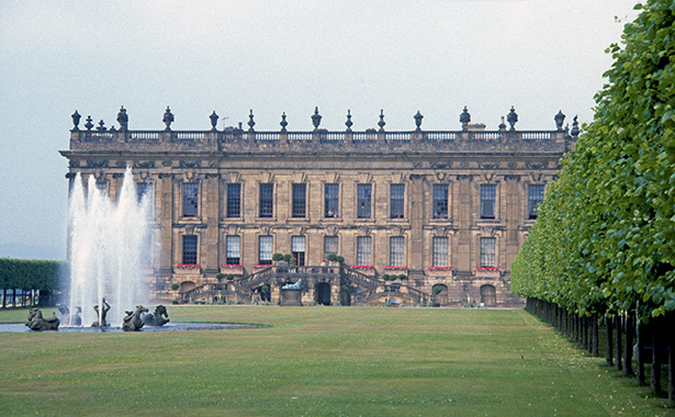 Chatsworth House in England photographed in 1984.  There is a lawn with fountains in the lower third of the image, with a straight line of trees on the right, and the large stone facade of Chatsworth House takes up the middle horizontal bar of the photograph, with a blue-grey sky at the top of the image.