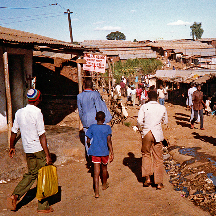 People walk down a dirt street with trash and sewage in teh middle of the street; a health clinic sign appears in the middle distance