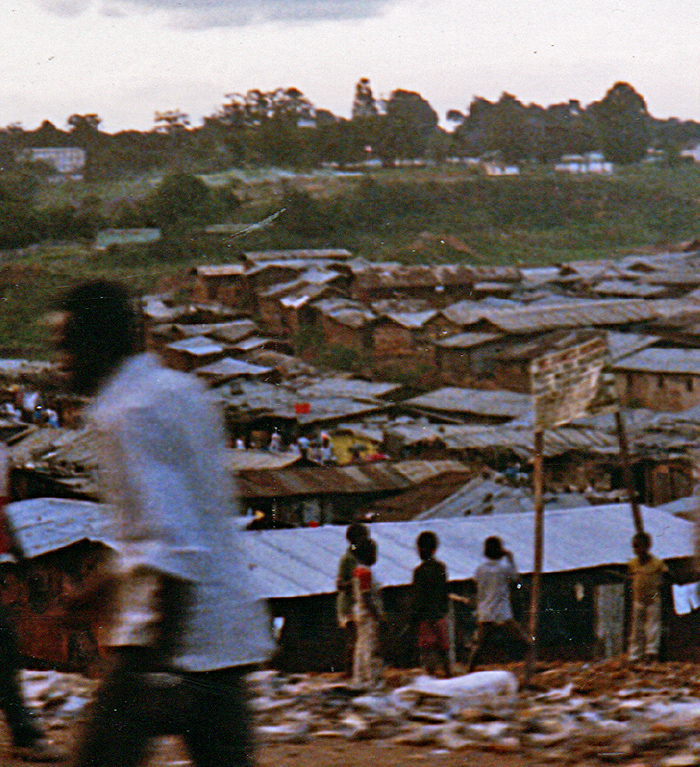 People walk along the street with informal housing in the background and garbage by the street