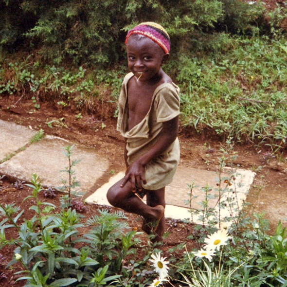 A young boy with a straw in his mouth stands on one leg and smiles