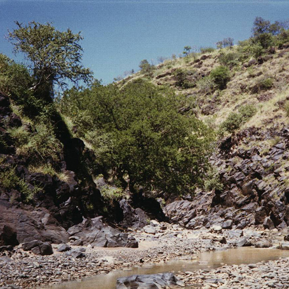 Where the creek came out of the ravine at Kositei Mission a muddy small creek, shallow and narrow, flows among many smooth stones with a bluff as a backdrop