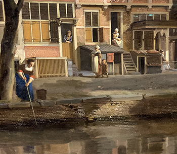 A woman pokes at the canal with a long pole, while behind her people look out from their doors as a woman with a child pass by on the street.