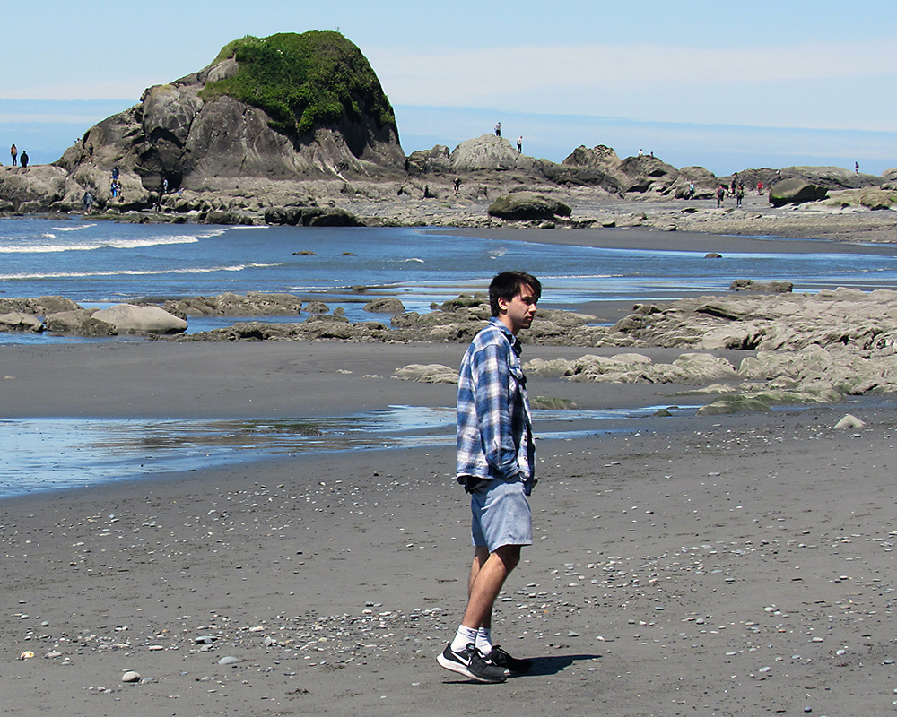 Arthur stands on the beach with rocks and the ocean behind him