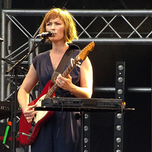 Mina Tindle up on stage singing "Pas les saisons", she is squinting into the lights to see the crowd, holding her guitar, with her mouth near the standing microphone.
